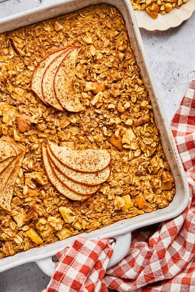 A close up of baked oatmeal with apples in a white baking dish. Around the pan is a red and white checkered kitchen towel and a plate of walnuts on it.