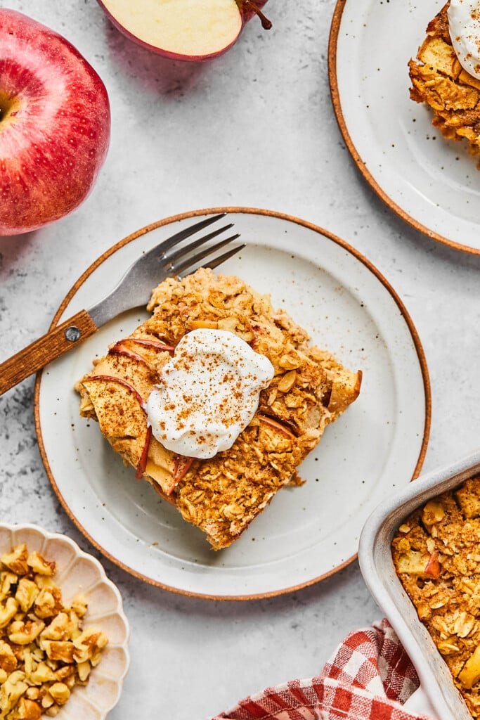 A piece of apple baked oatmeal on a plate with a fork on it. The oatmeal is topped with whipped cream and cinnamon. Around the plate is the pan of the rest of the baked oatmeal, a red and white checkered kitchen towel, a bowl of walnuts, a whole red apple and an apple that has been cut in half, and another plate with a piece of oatmeal on it.