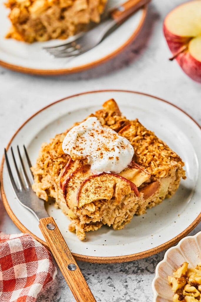 A piece of baked apple oatmeal on a plate. It is garnished with greek yogurt and cinnamon and a fork is also on the plate. Around the plate is a bowl of walnuts, a red and white checkered kitchen towel, another plate with a piece of apple oatmeal, and half a red apple.