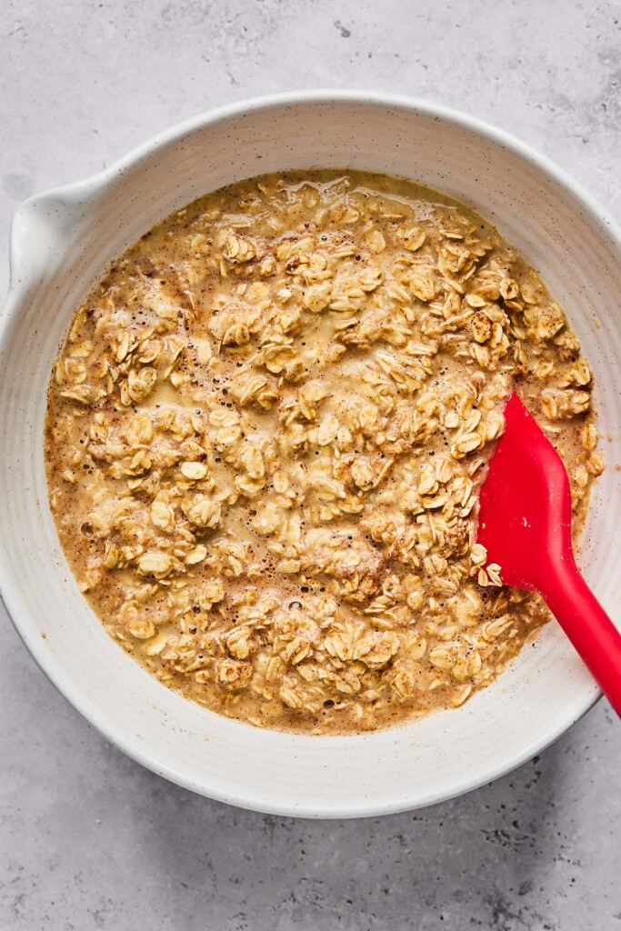 A mixing bowl filled with a batter of milk, eggs, rolled oats, and spices. A red spatula is also in the bowl.