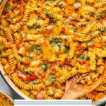 One pot enchilada pasta with ground beef, veggies, and melted cheese in a large enameled pan. It is garnished with cilantro and there is wooden spoon in the pan. Around the pan is a tan kitchen towel and a bowl of shredded cheese.
