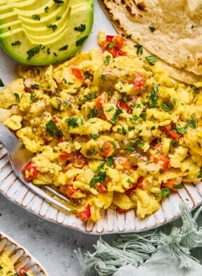 A plate of migas, sliced avocado, and tortillas. The migas are garnished with salsa and cilantro. Next to the plate is a green kitchen towel and another plate of migas.