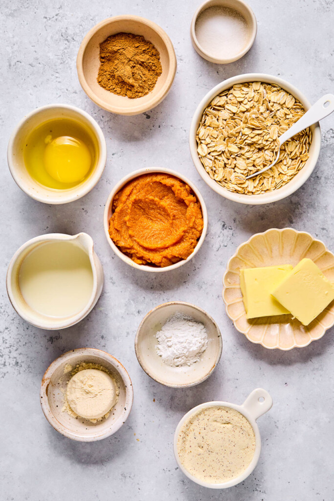 A small dish of salt, a bowl of rolled oats with a spoon in it, a plate with two slices of butter, a bowl of baking powder, a bowl of vanilla protein powder, a small bowl of coconut flour, a small pitcher of milk, a bowl of pumpkin puree, a bowl with a cracked egg in it, and a small bowl of pumpkin spice on a grey concrete counter.