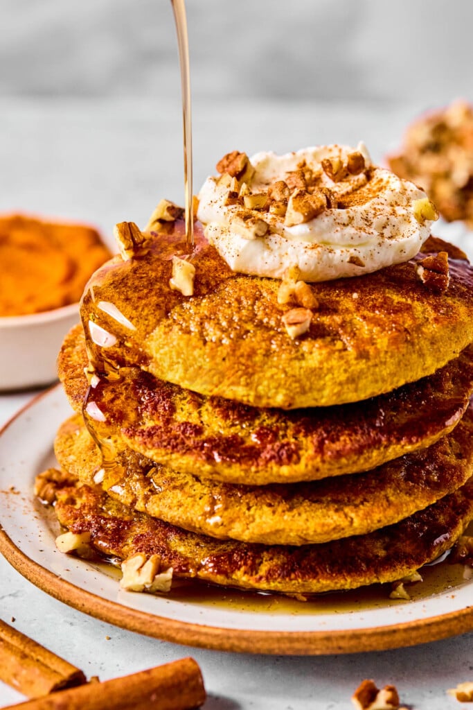 A plate with a stack of protein pumpkin pancakes topped with greek yogurt, pecan pieces, and cinnamon with maple syrup being poured on them. Around the plate are some cinnamon sticks, some pecan pieces, and a bowl of pumpkin puree.