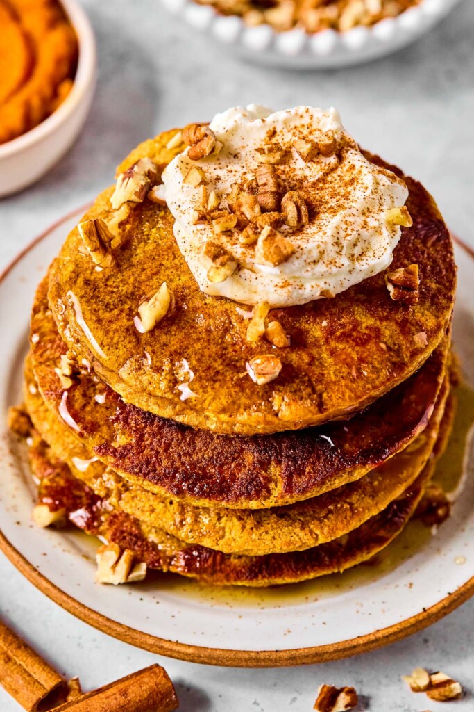 A stack of pumpkin protein pancakes topped with greek yogurt, pecan pieces, cinnamon, and maple syrup on a plate. Around the plate are some cinnamon sticks, pecan pieces, a bowl of pumpkin puree, and a bowl of pecans.