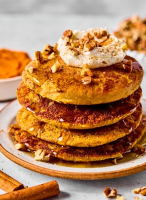 A stack of pumpkin protein pancakes topped with greek yogurt, pecan pieces, and maple syrup on a plate. Around the plate are some pecan pieces, cinnamon sticks, a bowl of pumpkin puree, and bowl of pecans.