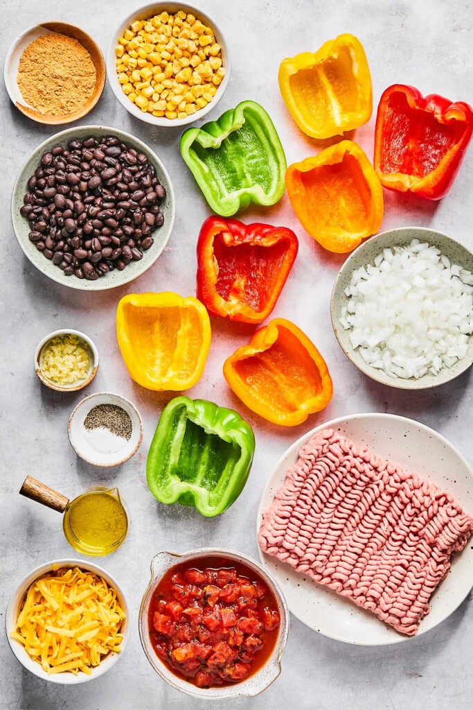 A variety of colored bell peppers that have been halved, a bowl of diced yellow onion, a plate of raw ground turkey, a bowl of diced fire roasted tomatoes, a bowl of shredded cheese, a small cup of olive oil, a small bowl of salt and pepper, a small bowl of minced garlic, a bowl of black beans, a bowl of taco seasoning, and a bowl of corn on a grey concrete countertop.