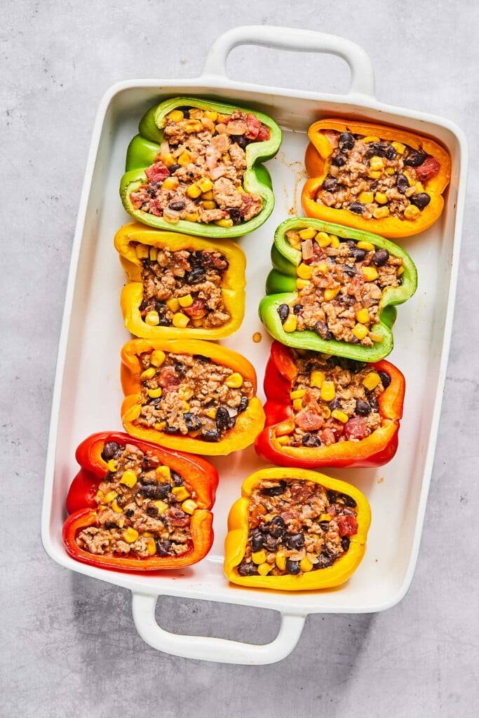 Par-baked halved bell peppers stuffed with a ground turkey, bean, and corn mixture in a large white casserole dish.