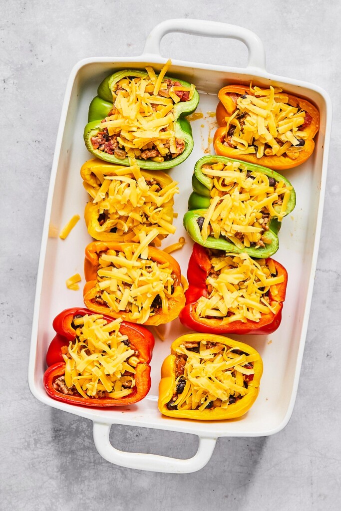 Par-baked halved bell peppers stuffed with a ground turkey, bean, and corn mixture with shredded cheese on top in a large white casserole dish.