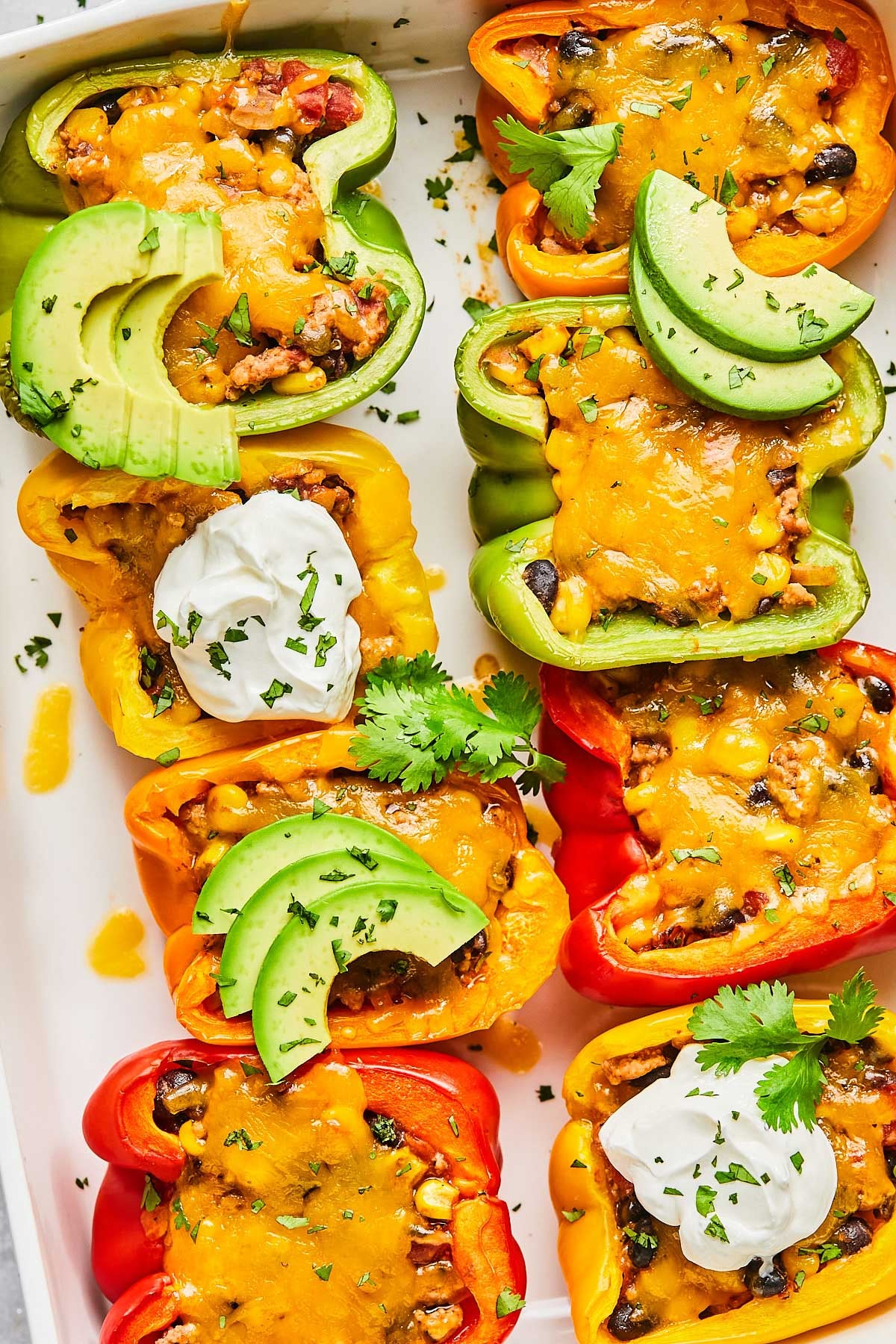 An overhead shot of taco stuffed peppers with melted cheese topped with sour cream, avocado slices, and cilantro in a white casserole dish.