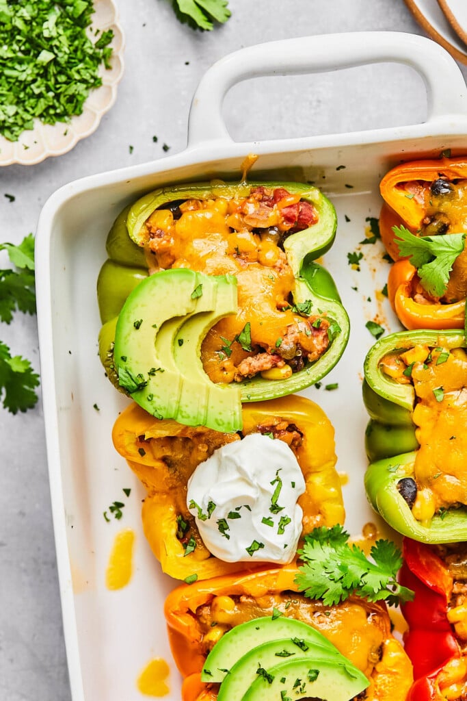 Mexican stuffed peppers with ground turkey and melted cheese in a large white casserole dish. They are garnished with sour cream, sliced avocado, and cilantro. Next to the pan is some cilantro leave and a plate of chopped cilantro.