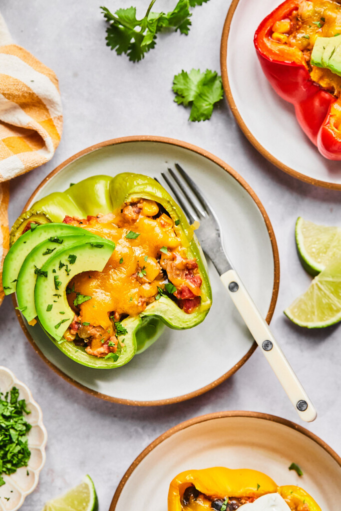 Healthy stuffed pepper with melted cheese and topped with sliced avocado and cilantro on a white plate. Around the plate is two more plates with stuffed peppers on them, a plate with cilantro, some lime wedges, and a striped kitchen towel.