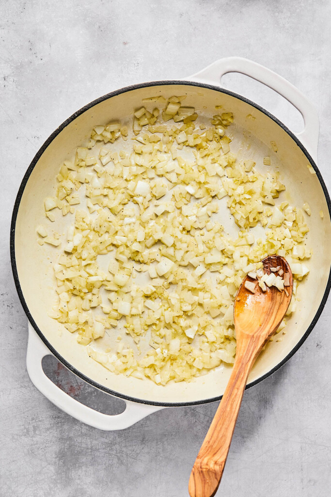 Large enameled white pan filled with sautéed onions and garlic. There is a wooden spoon in the pan as well.