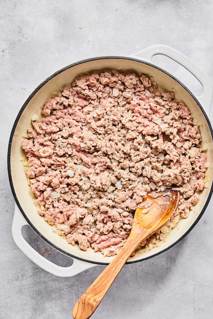 Large enameled white pan with ground turkey being cooked in it. There is a wooden spoon in the pan as well.