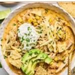 A bowl of healthy white chicken chili topped with greek yogurt, sliced avocado, shredded cheese, tortilla chips, and cilantro. There is a spoon in the bowl as well.