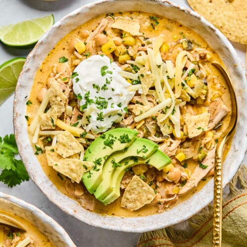 A bowl of healthy white chicken chili topped with greek yogurt, sliced avocado, shredded cheese, tortilla chips, and cilantro. There is a spoon in the bowl as well. Around the bowl are some tortilla chips, a tan kitchen towel, another bowl of spoon, some lime wedges, and a plate of chopped cilantro.