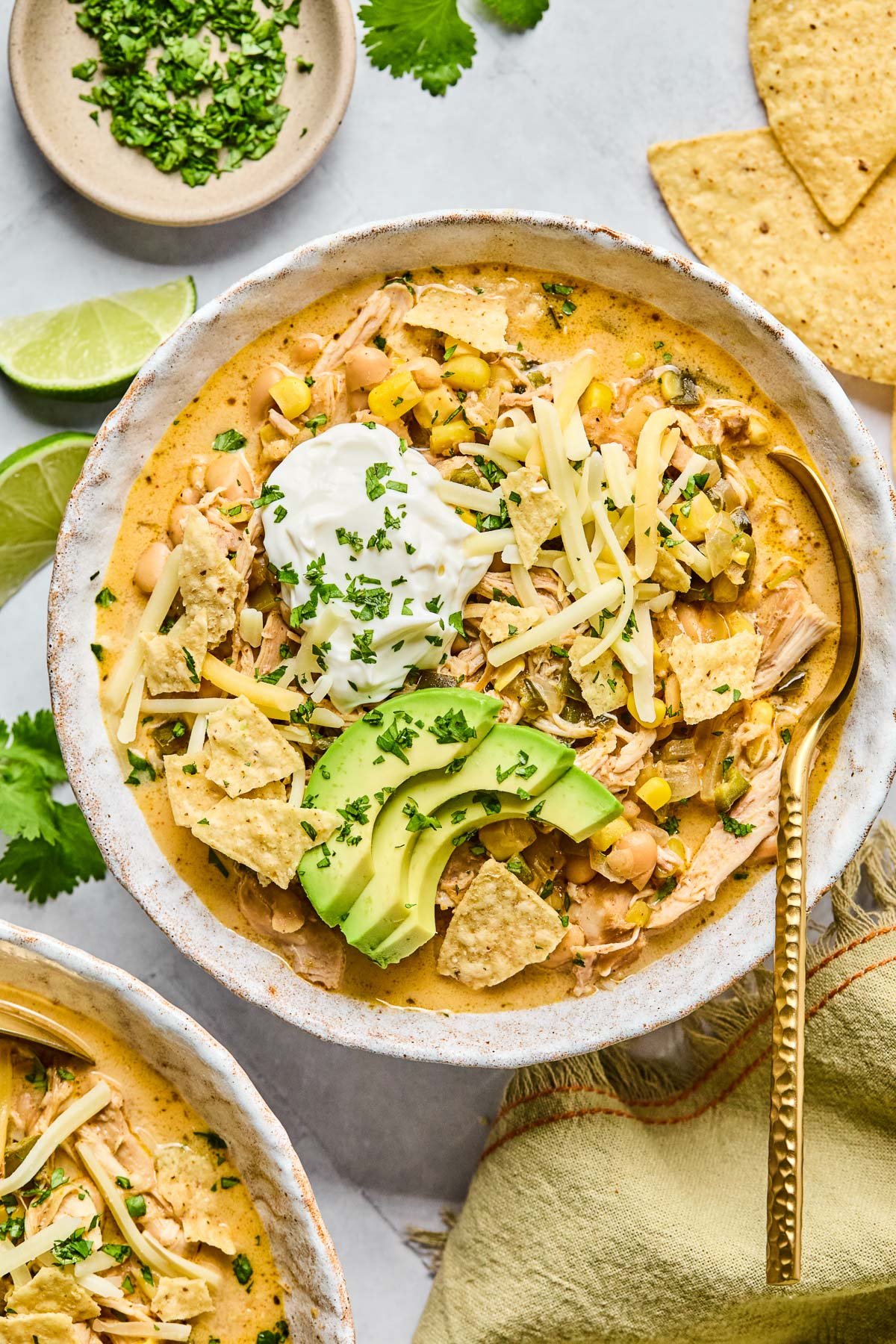 A bowl of healthy white chicken chili topped with greek yogurt, sliced avocado, shredded cheese, tortilla chips, and cilantro. There is a spoon in the bowl as well. Around the bowl are some tortilla chips, a tan kitchen towel, another bowl of spoon, some lime wedges, and a plate of chopped cilantro.