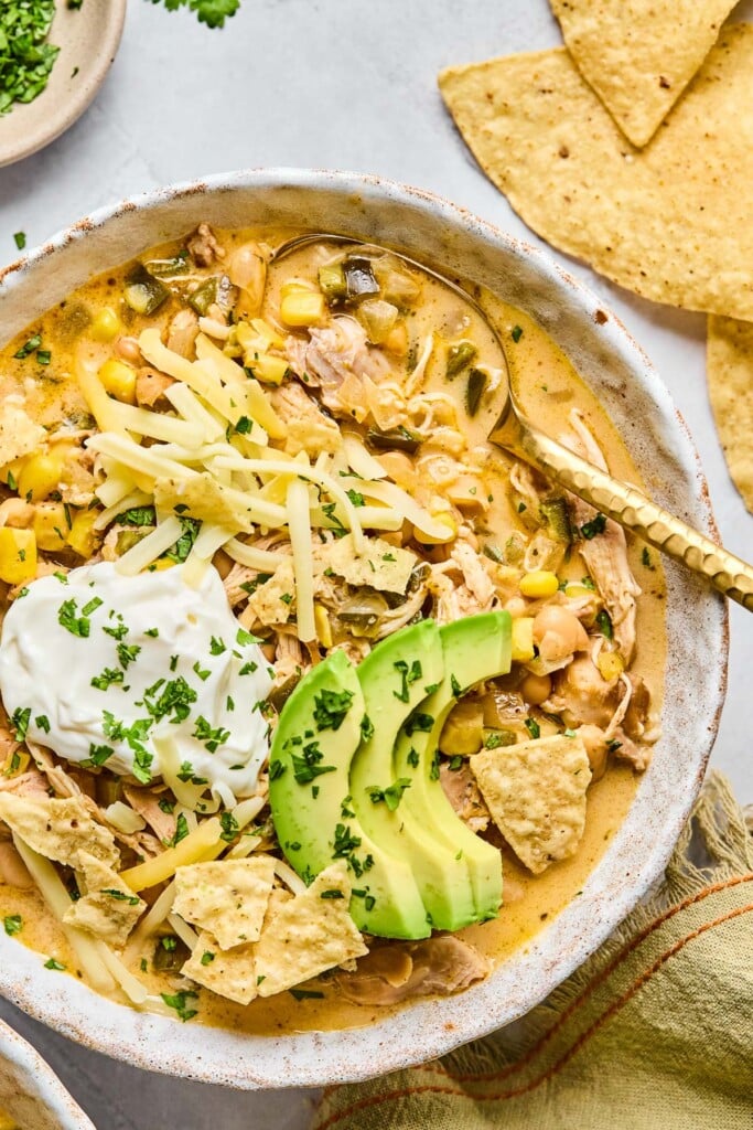 Crockpot white chicken chili in a bowl topped with greek yogurt, sliced avocado, shredded cheese, tortilla chips, and cilantro. There is a gold spoon in the bowl as well. Around the bowl are some tortilla chips, a tan kitchen towel, and a plate of chopped cilantro.