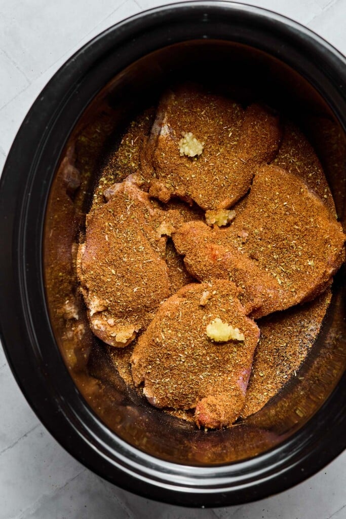 Slow cooker with chicken thighs covered with spices in it. There is also some minced garlic on top of the seasoned chicken.