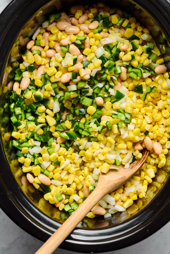 Overhead shot of a slow cooker filled with mixture of white beans, canned corn, diced onion, canned green chiles, and diced poblano pepper, and broth. There is a wooden spoon in the pot as well.