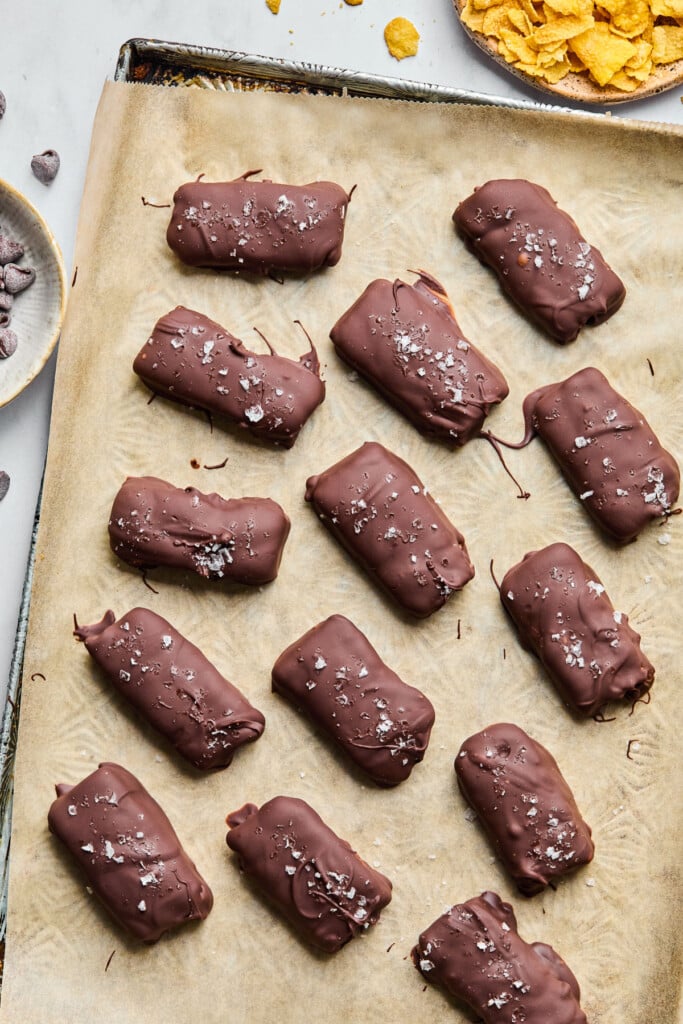 Healthy butterfinger candy bars on a baking sheet lined with parchment paper. Next to the baking sheet is a bowl of chocolate chips and a bowl of corn flakes.