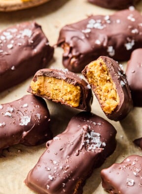 Homemade butterfinger candy bars on a piece of parchment paper. One of the bars is broken in half so you can see the inside of the bar. Off in the corner is a bowl of cornflakes.