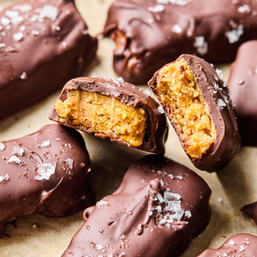 Homemade butterfinger candy bars on a piece of parchment paper. One of the bars is broken in half so you can see the inside of the bar. Off in the corner is a bowl of cornflakes.