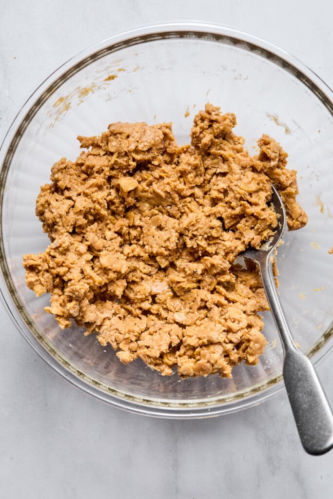 Peanut butter mixed with cornflakes and flour in glass mixing bowl with a spoon in it.