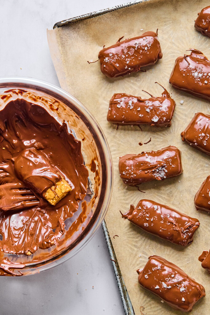 A baking sheet of candy bars that have just been dipped into melted chocolate. Next to the baking sheet is a bowl of melted chocolate with a candy bar on a fork that is halfway dipped in chocolate.