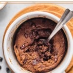 Overhead shot of a protein mug cake with a spoon in it with some of the middle scooped out. The mug is sitting on a small wooden coaster and around it are some mini chocolate chips.