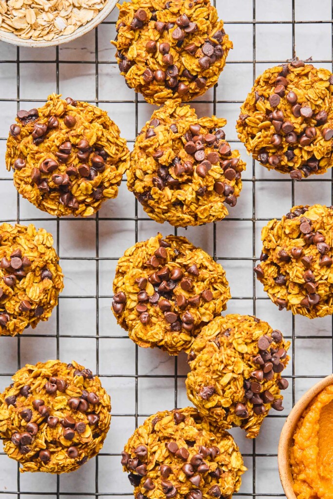 Overhead shot of oatmeal pumpkin muffins spread on a metal cooling rack. There is a bowl of pumpkin puree and a bowl of rolled oats on the rack as well.