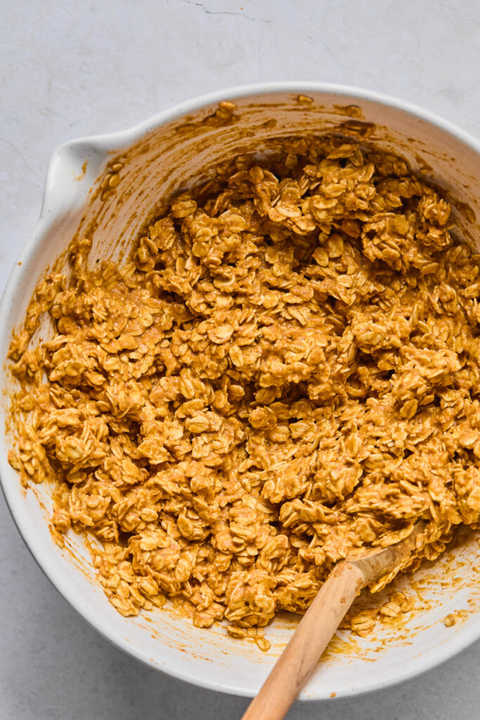 White mixing bowl with a wet batter with rolled oats in it. A wooden spoon is also in the bowl.