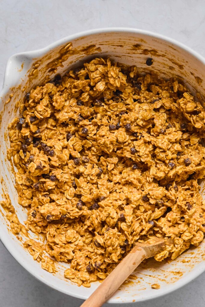 White mixing bowl with a pumpkin oatmeal batter with chocolate chips in it. A wooden spoon is also in the bowl.