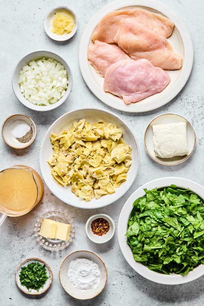 A plate of four raw chicken breasts, a plate of with a brick of cream cheese on it, a bowl of chopped spinach, a small bowl of flour, a small bowl of chopped parsley, a small bowl of red pepper flakes, a small plate of butter, a cup of bone broth, a small dish of salt and pepper, a bowl of diced onions, and a bowl of minced garlic on a grey counter.