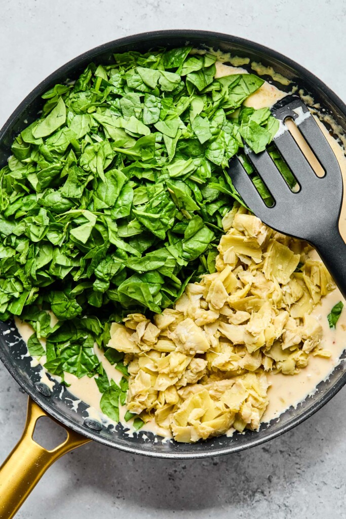 Skillet of a creamy white sauce with a bunch of fresh spinach and chopped artichokes overtop of it before it is mixed together.
