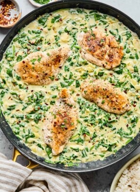 Spinach artichoke chicken skillet garnished with red pepper flakes. Next to the pan is a bowl of rice, a striped kitchen towel, a small bowl of red pepper flakes, and a small bowl of parsley.