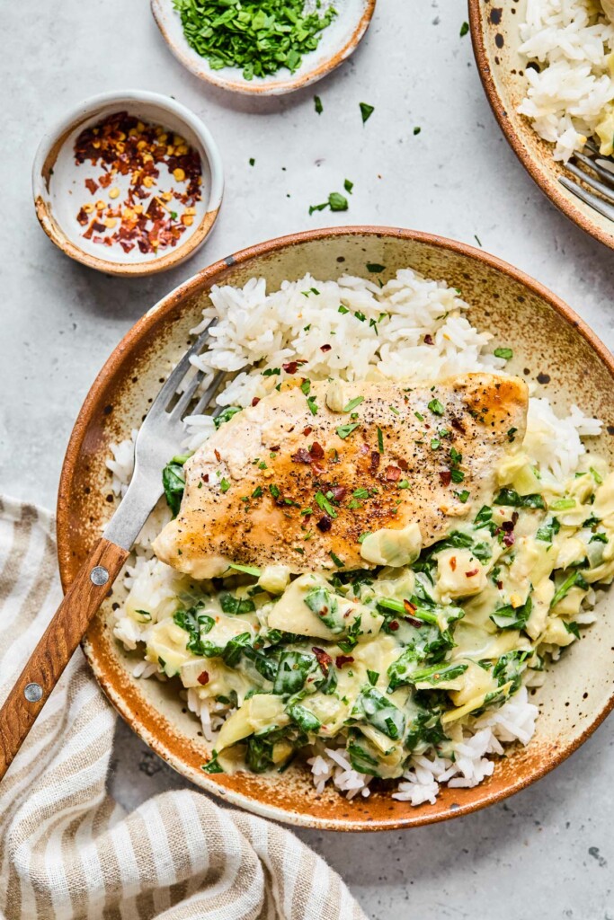 Chicken with spinach and artichokes in a creamy sauce over a bed of rice and garnished with red pepper flakes in a bowl. There is a fork in the bowl as well. Around the bowl is a striped kitchen towel, a small bowl of red pepper flakes, a small bowl of chopped parsley, and a bowl of rice.