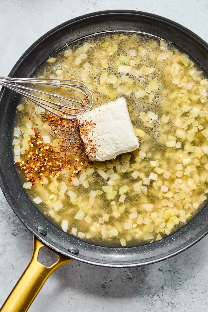 Overhead shot of a skillet with sautéed onions and garlic, bone broth, cream cheese, and red pepper flakes before it is mixed together.