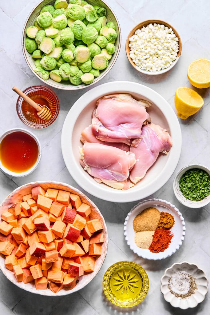 A bowl of crumbled goat cheese, a lemon cut in half, a shallow bowl of raw chicken thighs, a small bowl of chopped cilantro, a small plate of four different spices piled next to each other, a small dish of salt and pepper, a small cup of olive oil, a bowl of diced raw sweet potatoes, a bowl of hot honey, a bowl of honey with a honey dipper in it, and a bowl of halved raw Brussels sprouts on a grey counter.