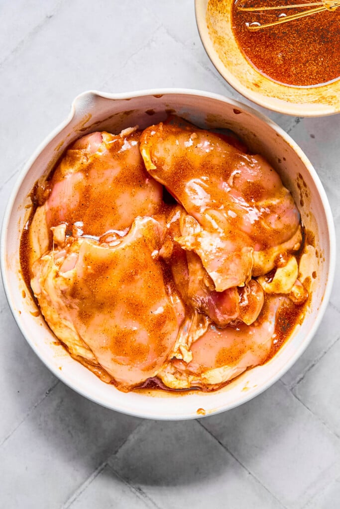Chicken thighs marinating in mixture of oil and spices a bowl. Next to the bowl is more marinade with a small whisk in it.