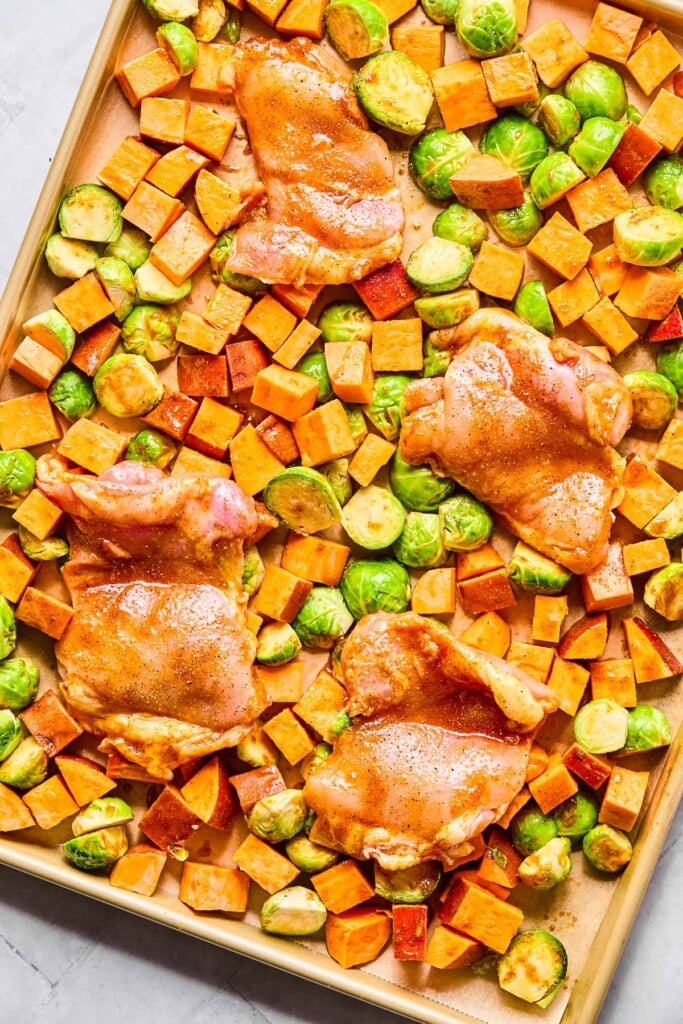 Parchment paper lined baking sheet with raw chicken thighs, diced sweet potatoes, and halved brussels sprouts all covered in oil and spices.