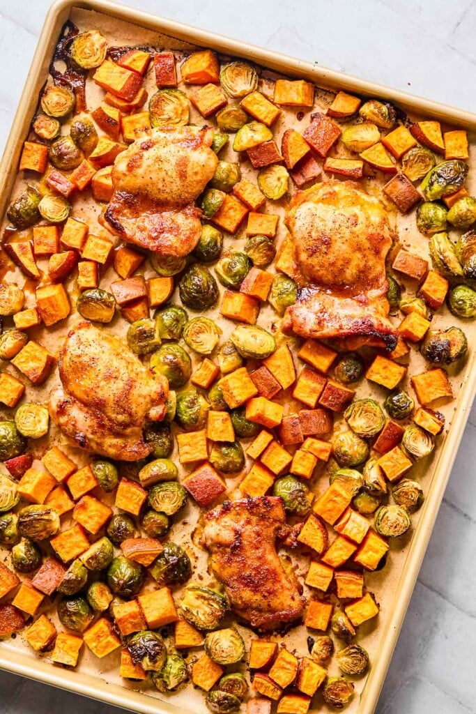 Parchment paper lined baking sheet with roasted seasoned chicken thighs, diced sweet potatoes, and halved brussels sprouts.