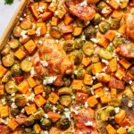 Sheet pan hot honey chicken with sweet potatoes and brussels sprouts topped with cilantro and goat cheese. A metal spatula is on the sheet pan with a few veggies on it.