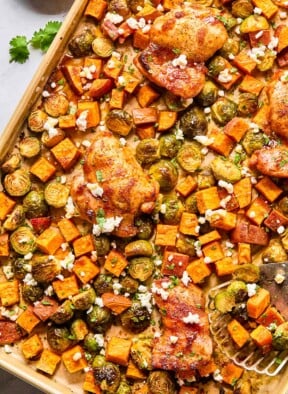 Sheet pan hot honey chicken with sweet potatoes and brussels sprouts topped with cilantro and goat cheese. A metal spatula is on the sheet pan with a few veggies on it.