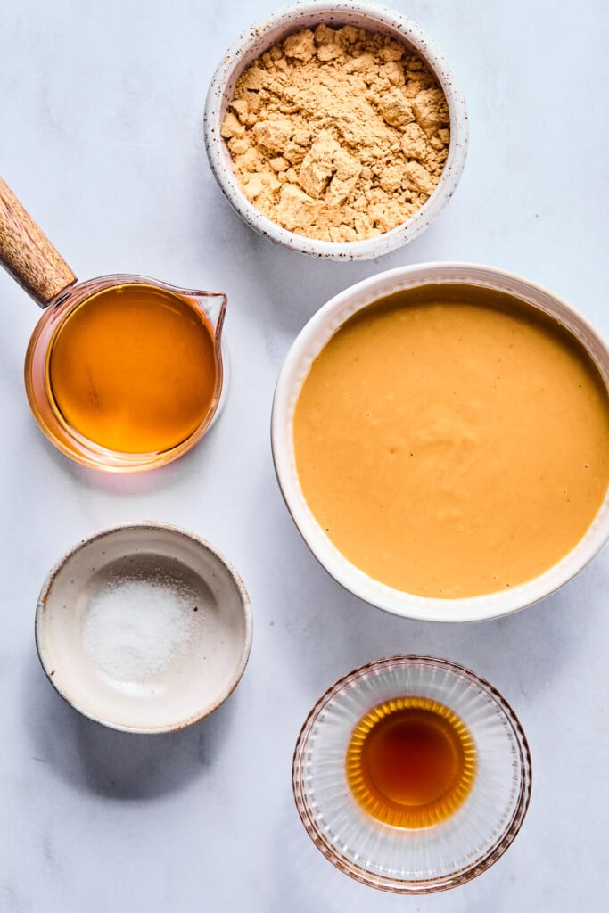 A bowl of flour, a bowl of creamy peanut butter, a bowl of vanilla extract, a bowl of salt, and a small cup of maple syrup on a grey counter.