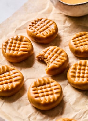 No bake peanut butter cookies on a piece of parchment paper. One of the cookies has a bite taken out of it. There is a bowl of peanut butter behind them.