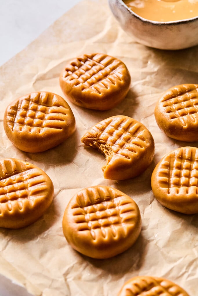 No bake peanut butter cookies on a piece of parchment paper. One of the cookies has a bite taken out of it. There is a bowl of peanut butter behind them.
