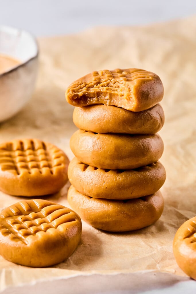 A stack of five peanut butter no bake cookies on parchment paper. The top one has a bite taken out of it. Around the stack are more cookies and a bowl of peanut butter.