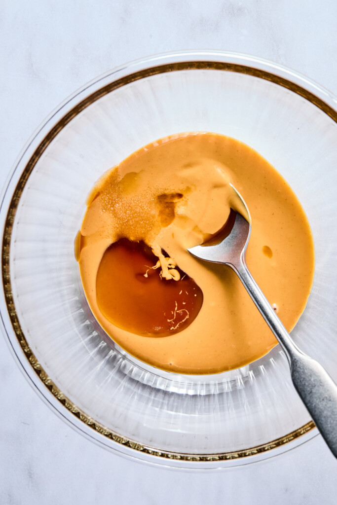A glass bowl with creamy peanut butter, maple syrup, and vanilla extract in it.