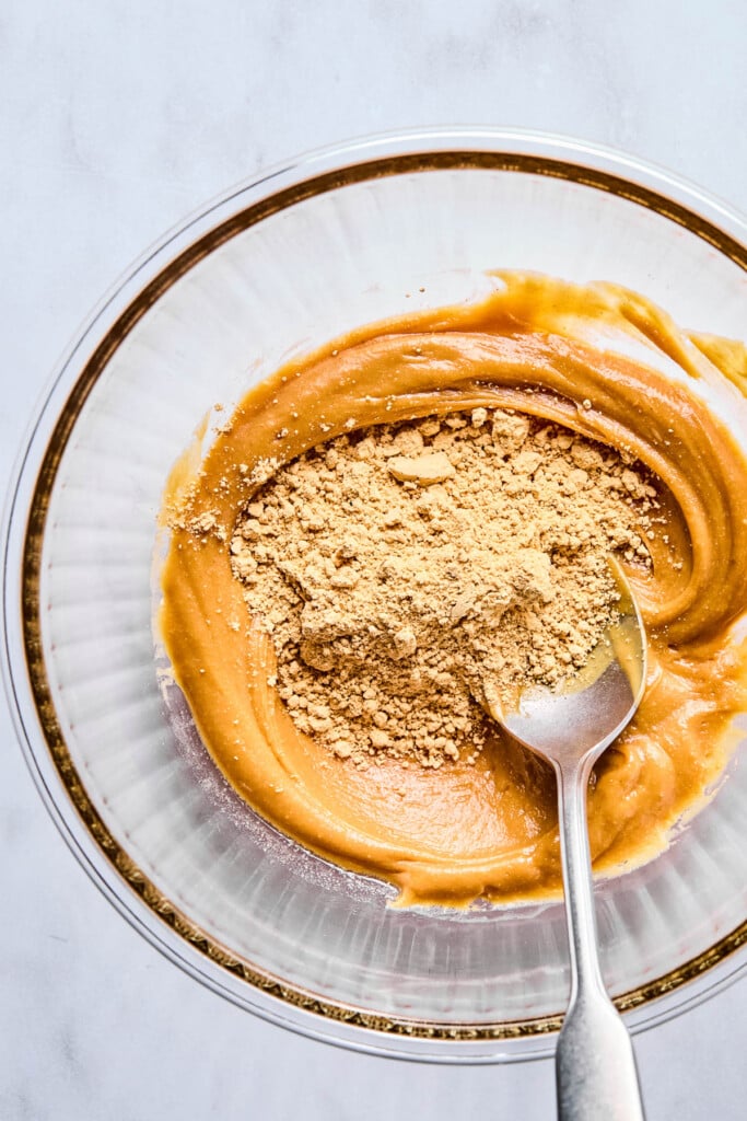 A bowl of creamy peanut butter batter with flour sprinkled on top before it is mixed together. A spoon is in the bowl too.